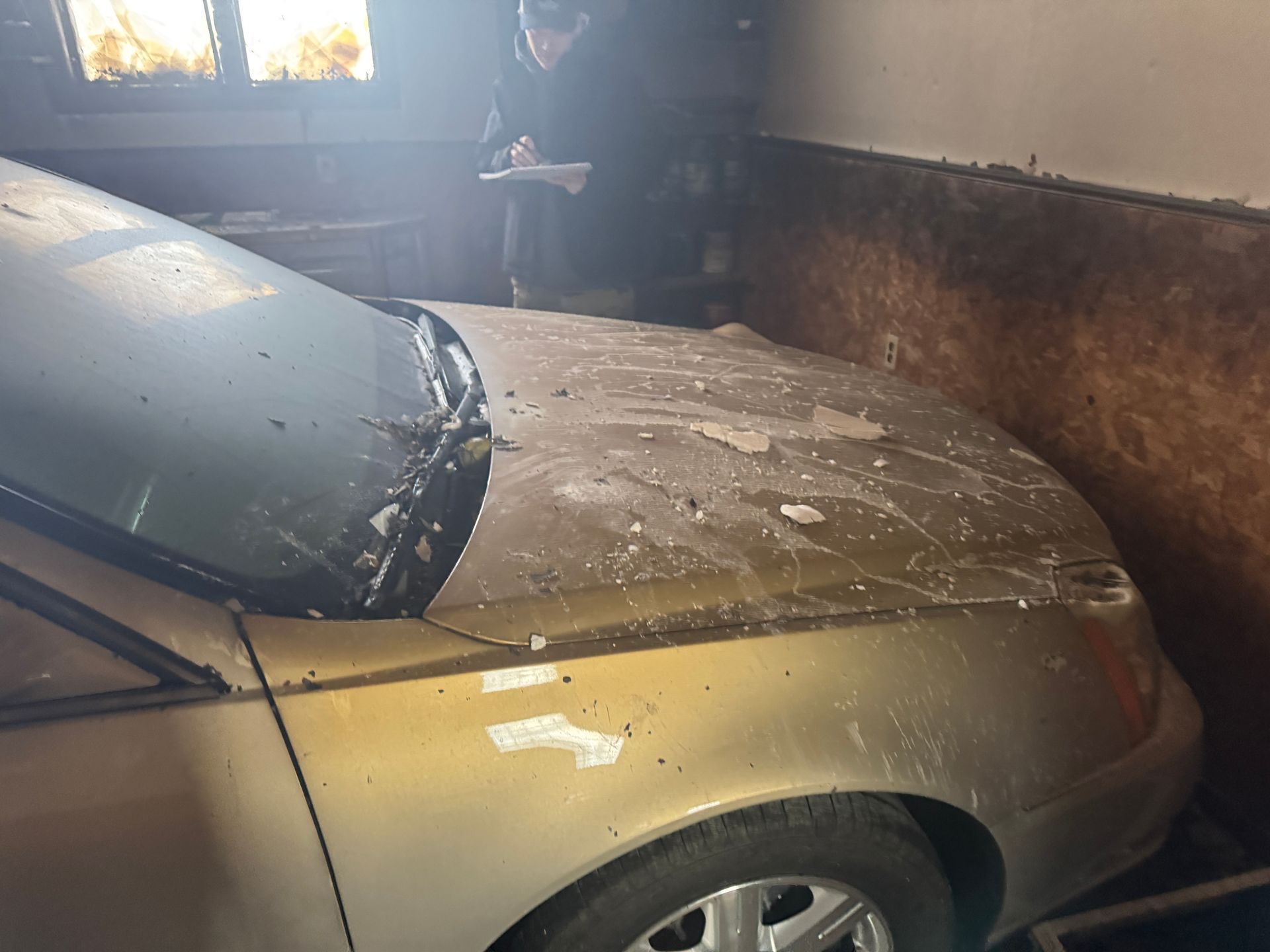 A gold car covered in dust sits in a garage; a person wearing a coat and hat is taking notes nearby.