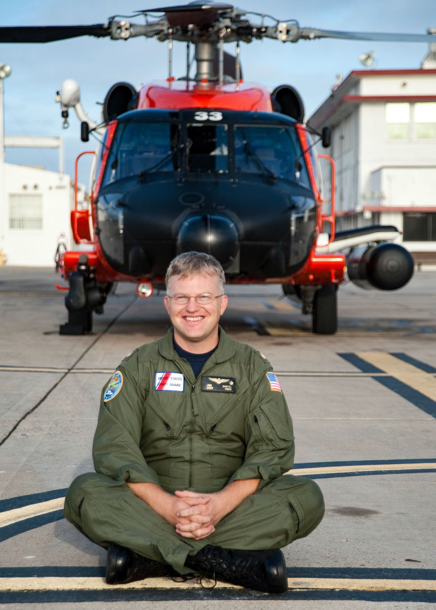Jon Bartel smiling and sitting on the ground in front of a Coast Guard helicopter