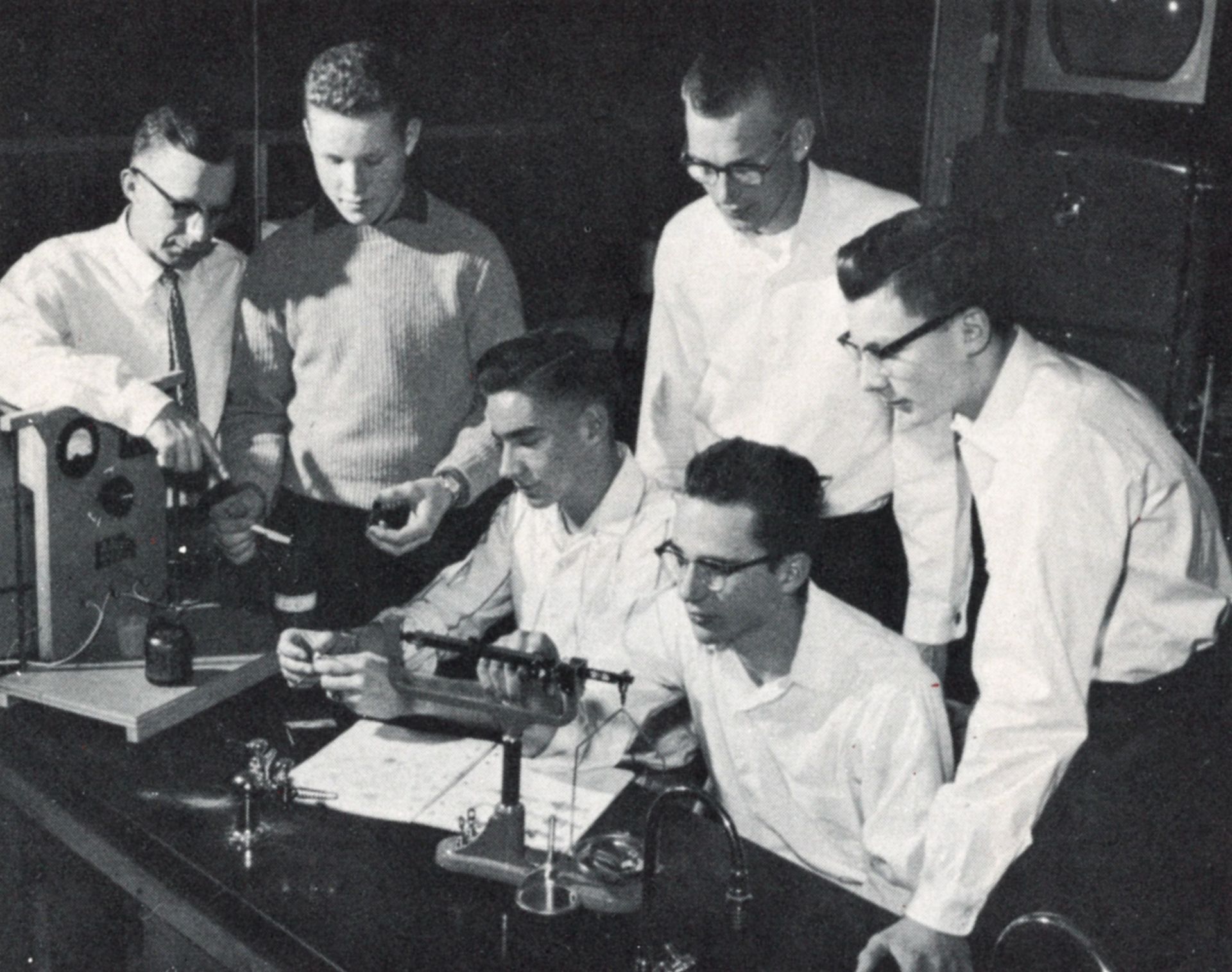 Six guys (one of whom is a teacher) gathered around a project on a science lab table.