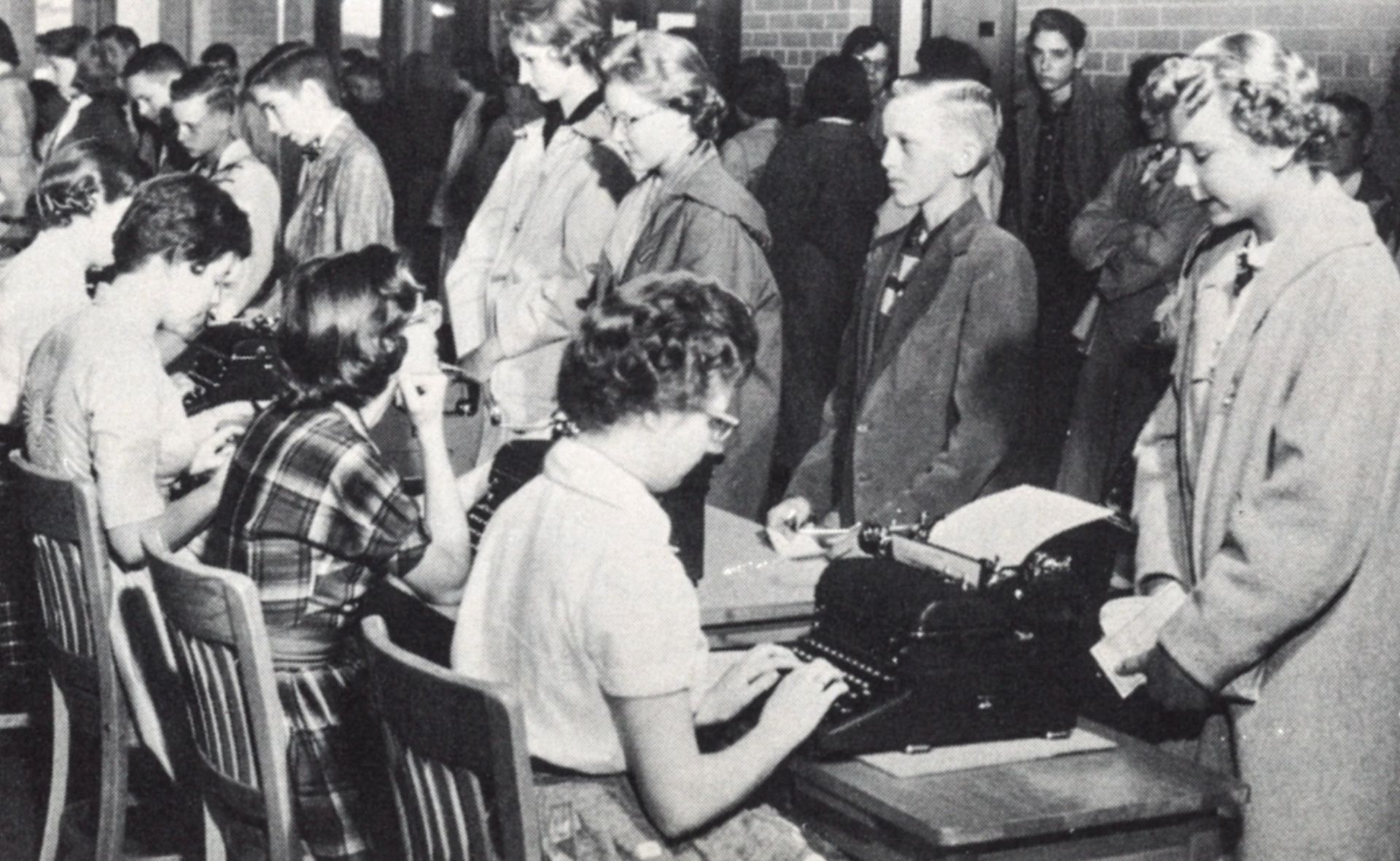 Row of women at typewriters with students lined up in front of them.