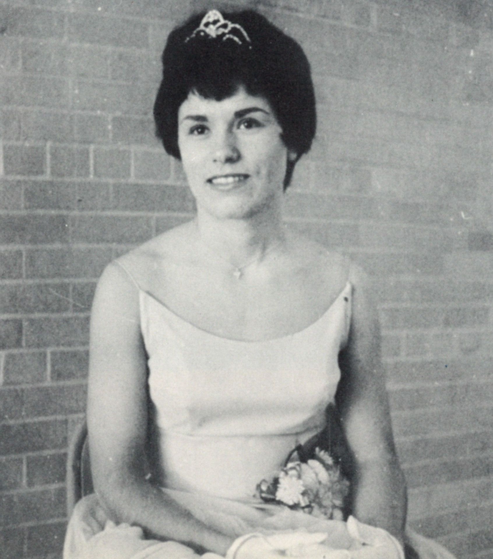 Homecoming Queen wearing gloves and crown, and with a flower corsage pinned at her waist, sitting in a chair