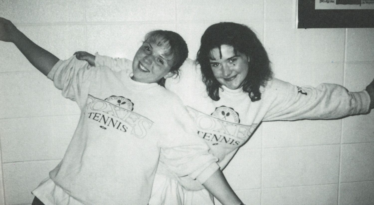 Two smiling girls wearing FVL Tennis sweatshirts, with their arms outstretched