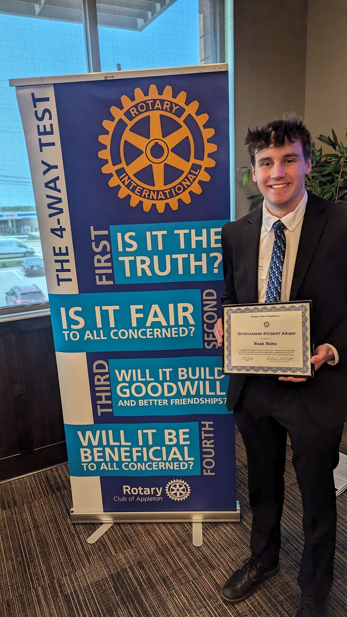 Noah Nolte holding his certificate, standing by a Rotary banner