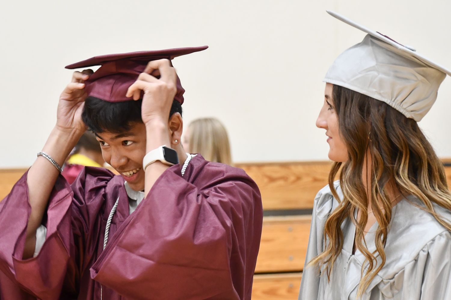 Guy and girl in the small gym prior to graduation. He is adjusting his mortar board and she is looking on, smiling
