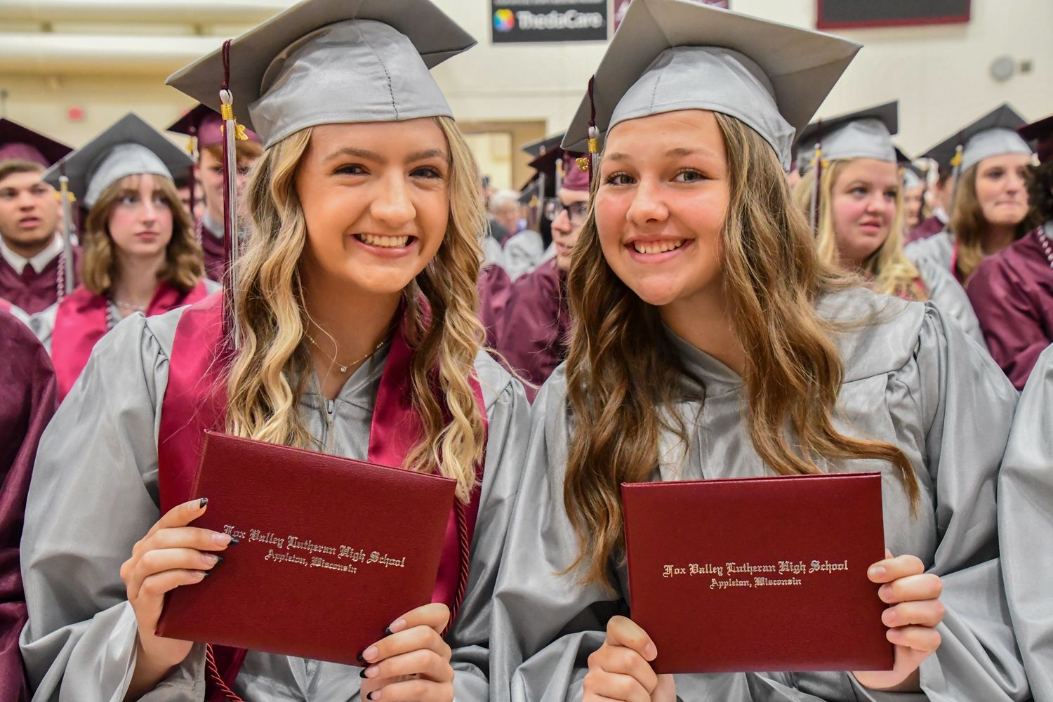 Two smiling girls, back in their seats holding up their diplomas