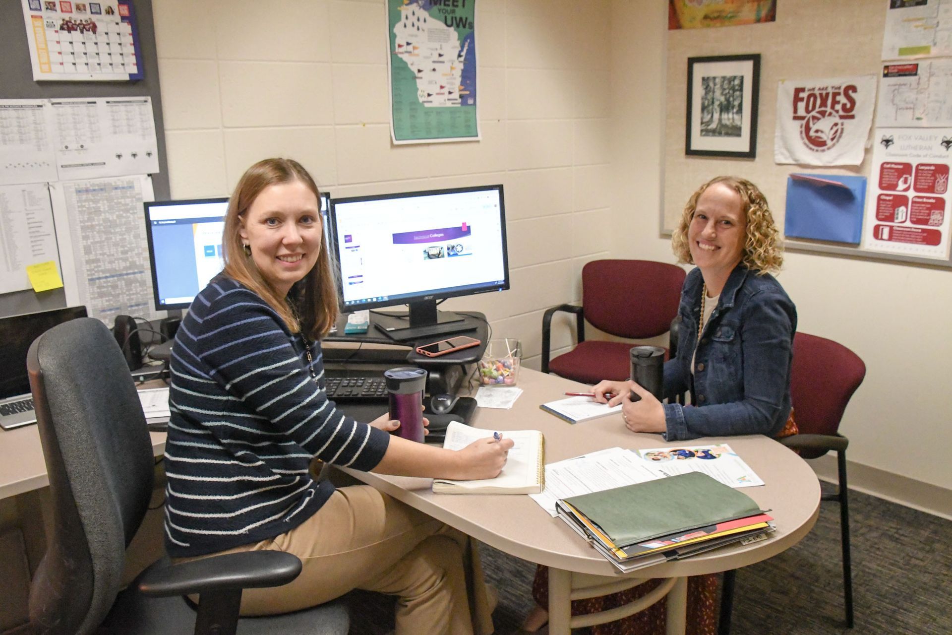 Aleay Friemark sitting at her desk with Heidi Wendland on the other side of the desk. Both of them are smiling and looking at the camera.
