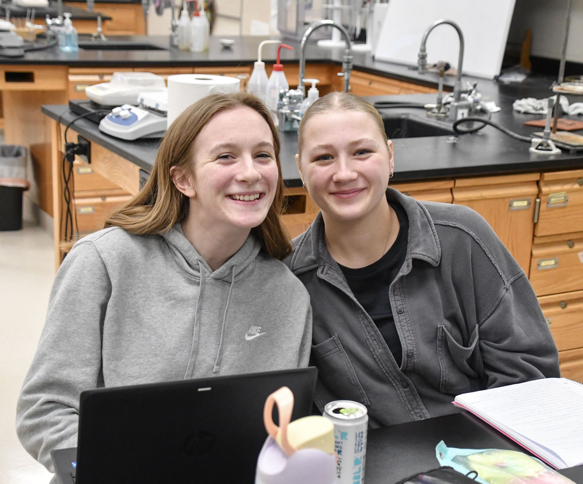 Two smiling girls sitting at table in science lab, smiling at the camera