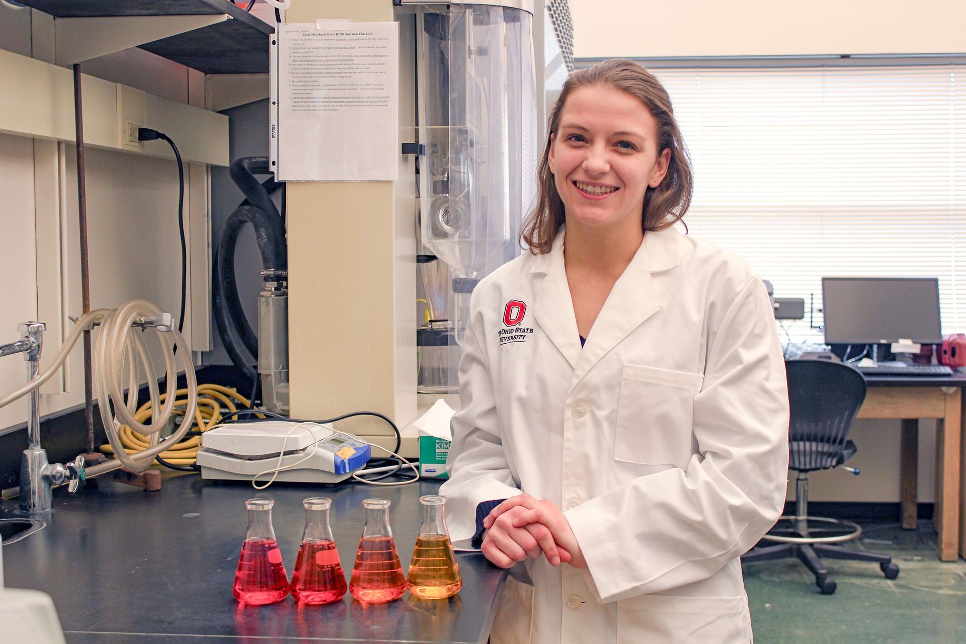 Danielle Voss wearing a lab coat, in a science lab, with 4 beakers on a table in front of her