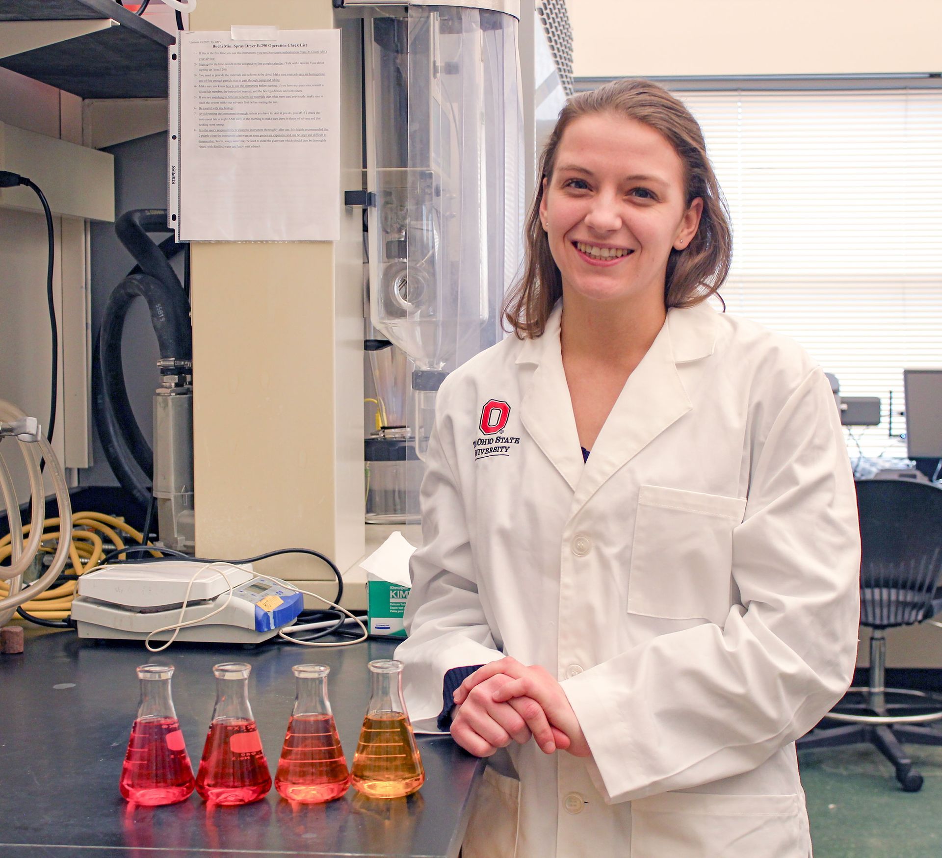 Smiling Danielle Voss in lab coat in a science lab with beakers on the table (partially filled with colorful liquids)