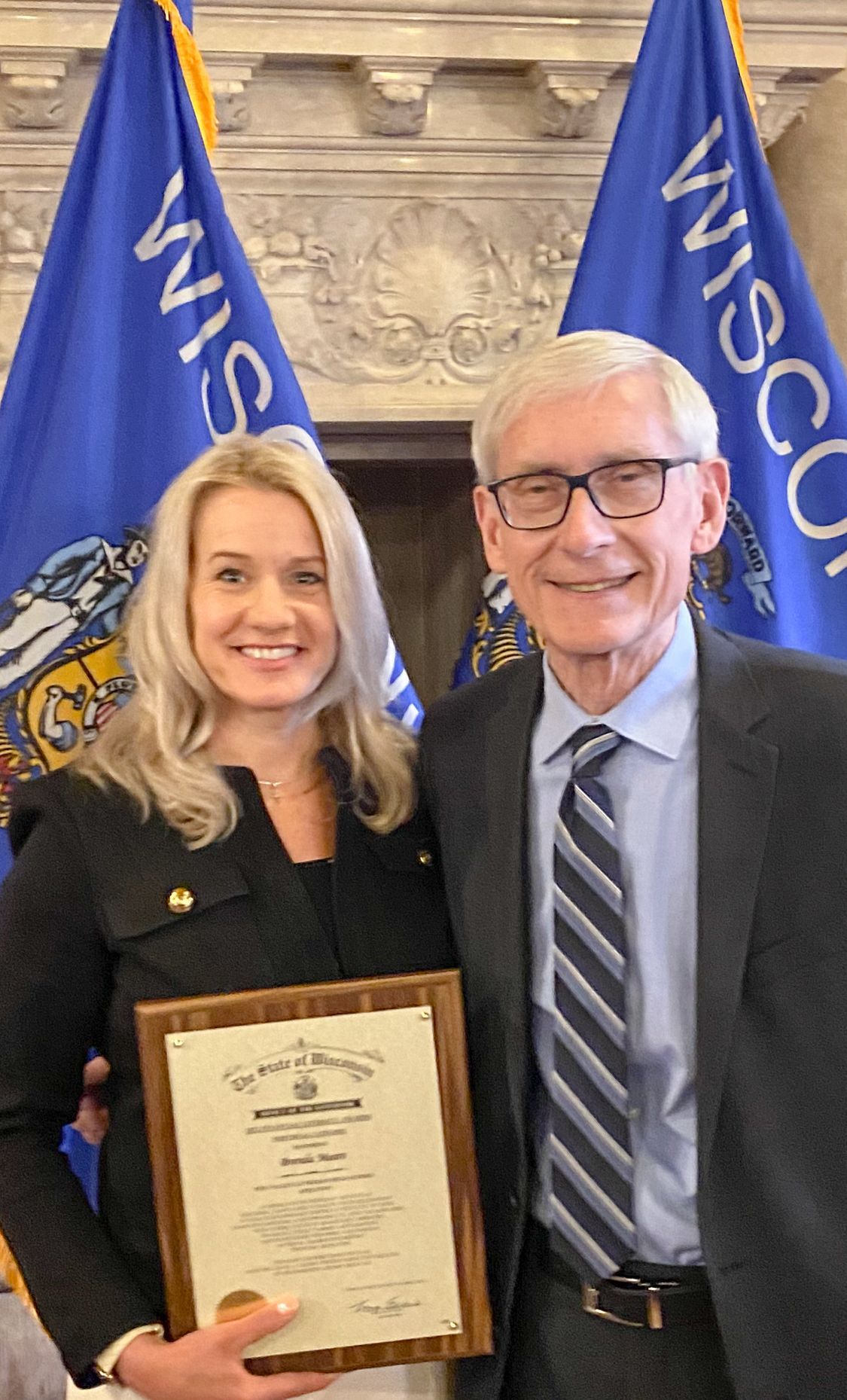Brenda Mears holding her plaque and standing next to Governor Evers with two Wisconsin flags behind them