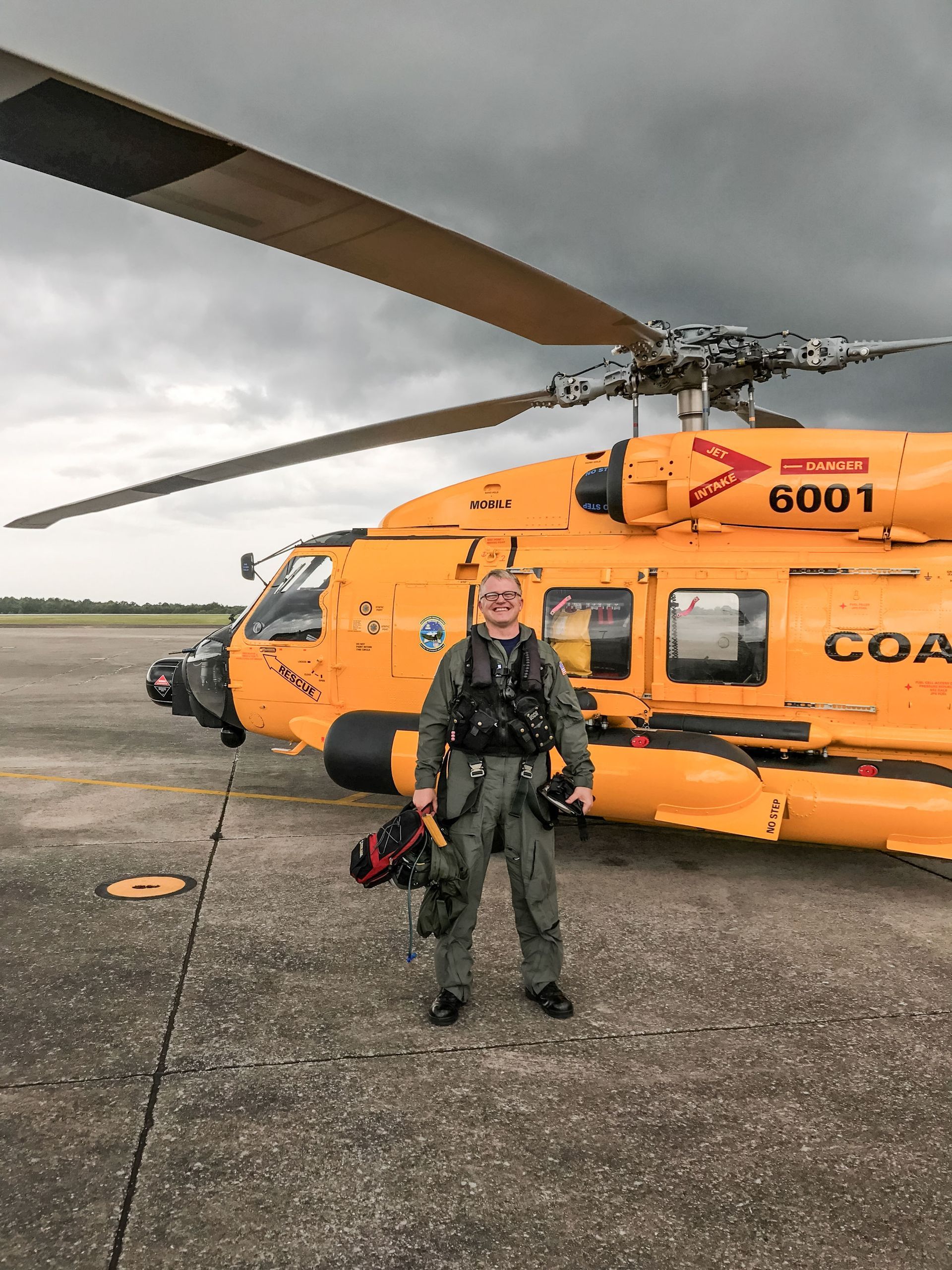Smiling Jon Bartel standing in front of a Coast Guard helicopter with cloudy skies above
