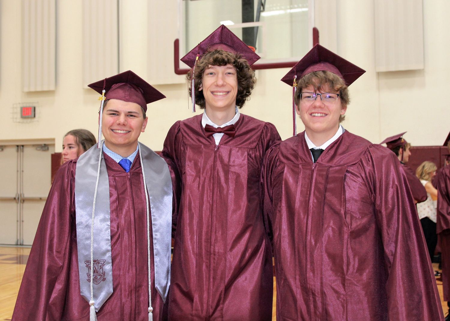 Three smiling guys in the small gym in cap and gown. Far left student also has a stole and cords