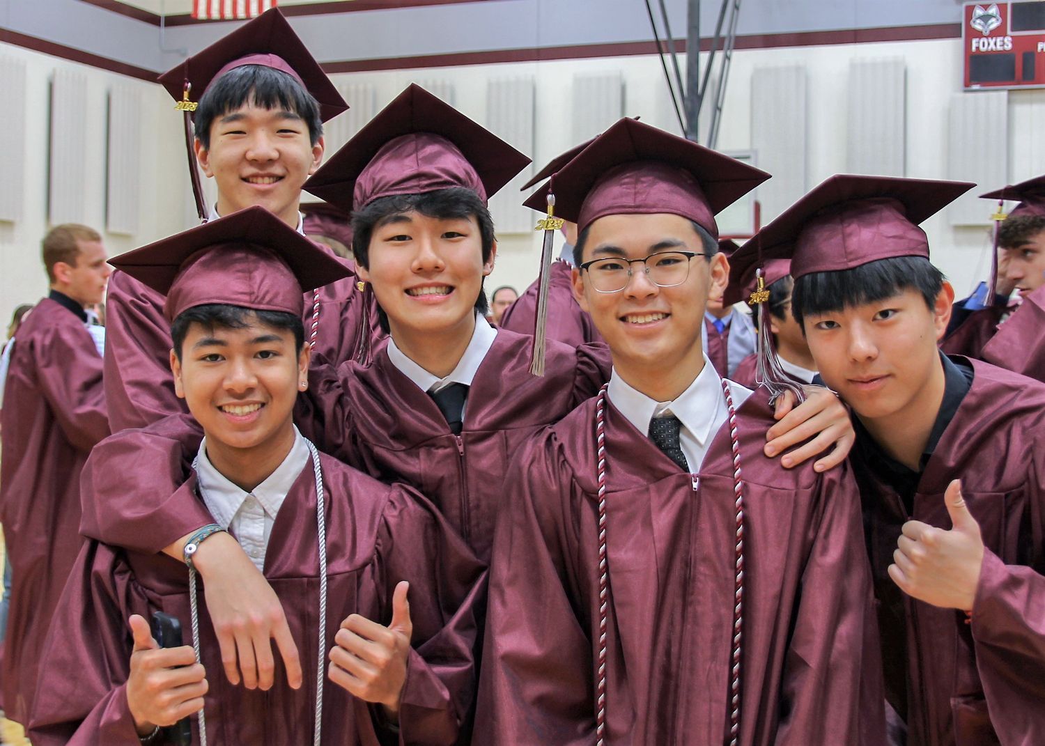 Group of 5 smiling guys, some with thumbs up, all wearing caps and gowns