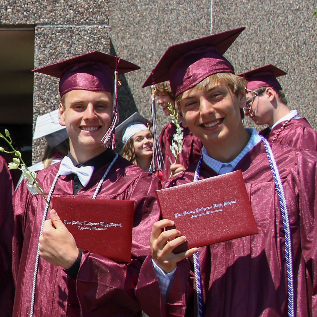 Two smiling guys, outside, holding up their diplomas