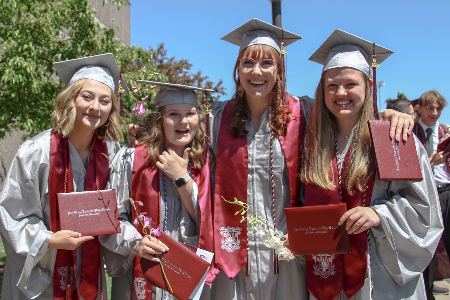 Four happy girls in caps gowns, and stoles, holding their diplomas outside