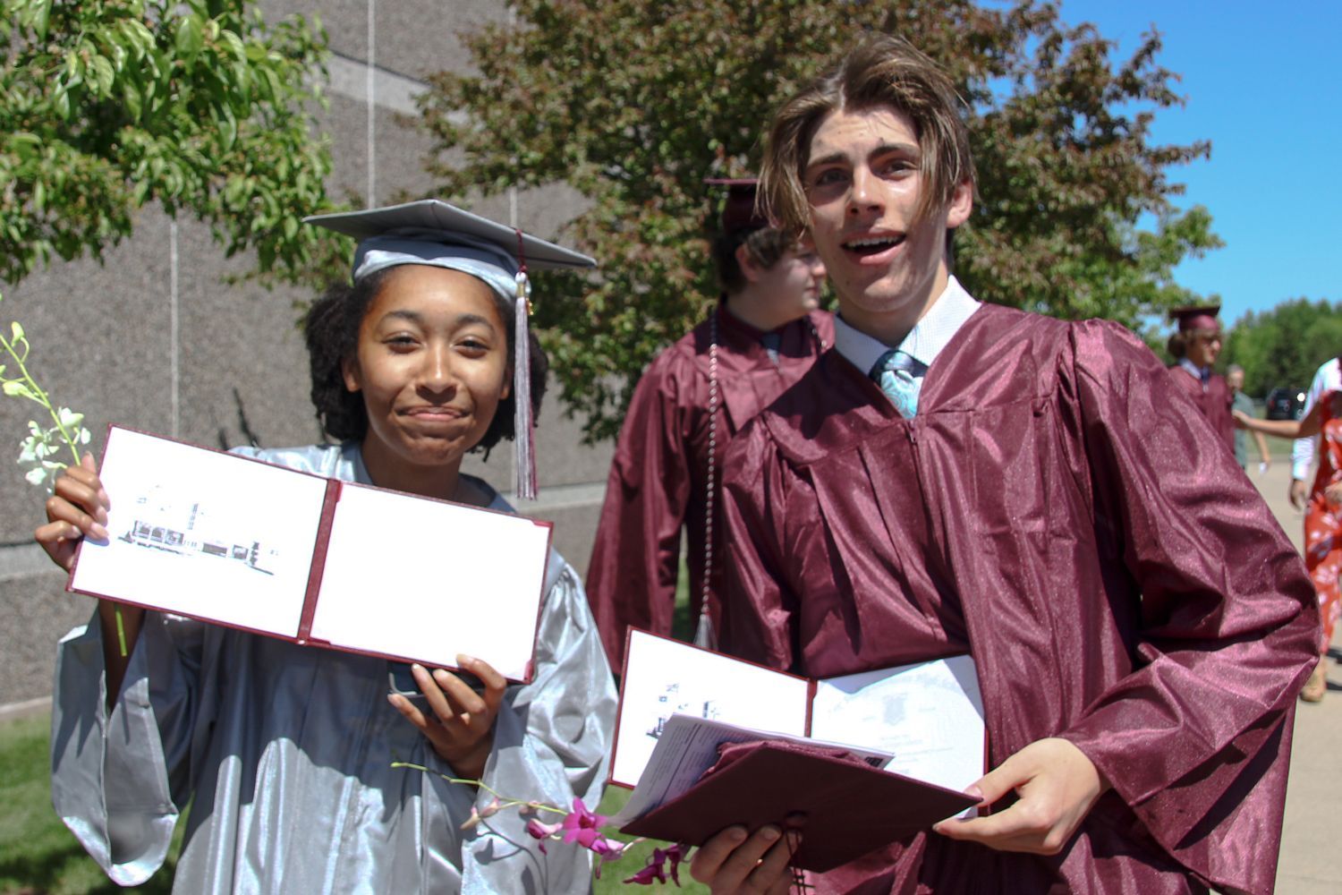 Smiling guy and girl, holding up their opened diplomas outside after the ceremony