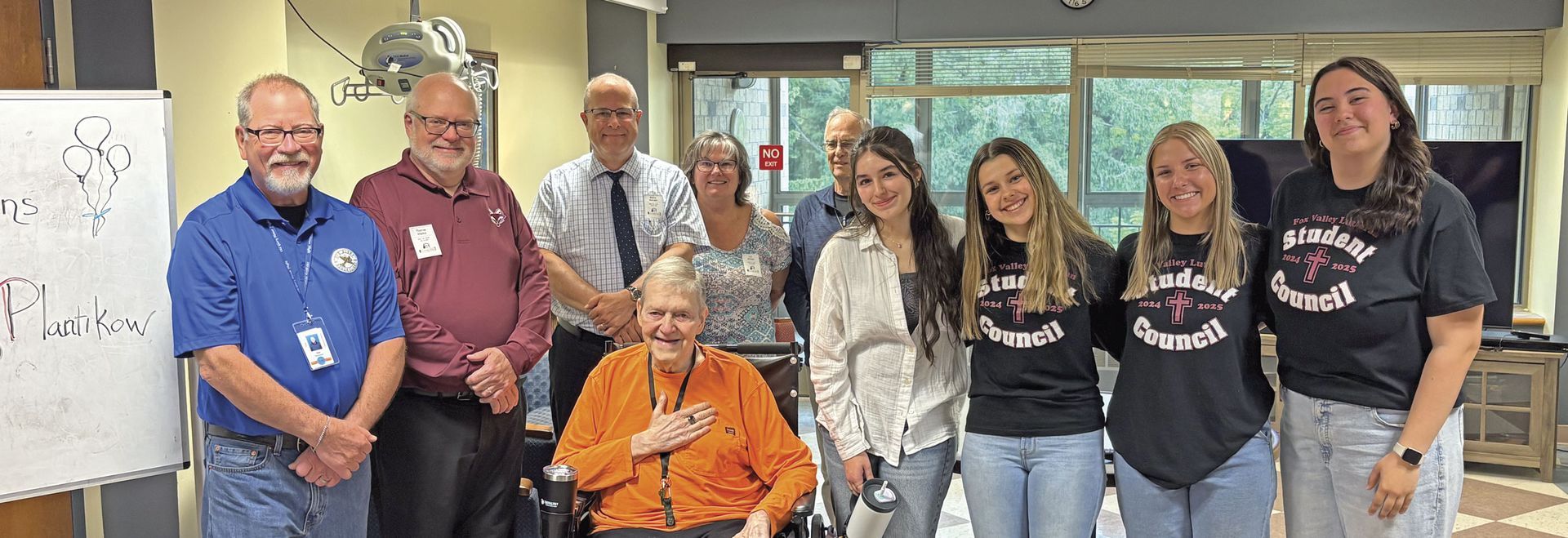 Nine smiling people standing around Jerry (in a wheelchair) at the nursing home. Jerry is displaying his new ring.