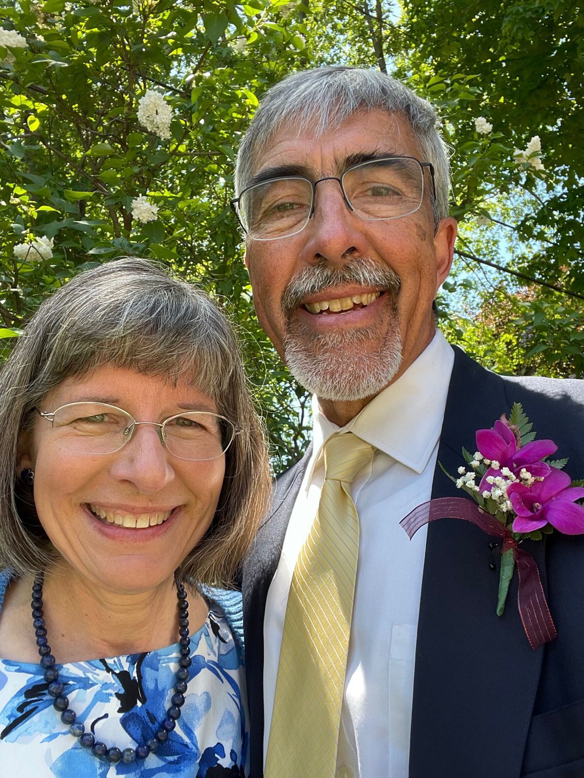 Pete Sehloff, with a purple flower on his suit lapel, with his wife, Sarah. They are outside with blossoming trees in the background