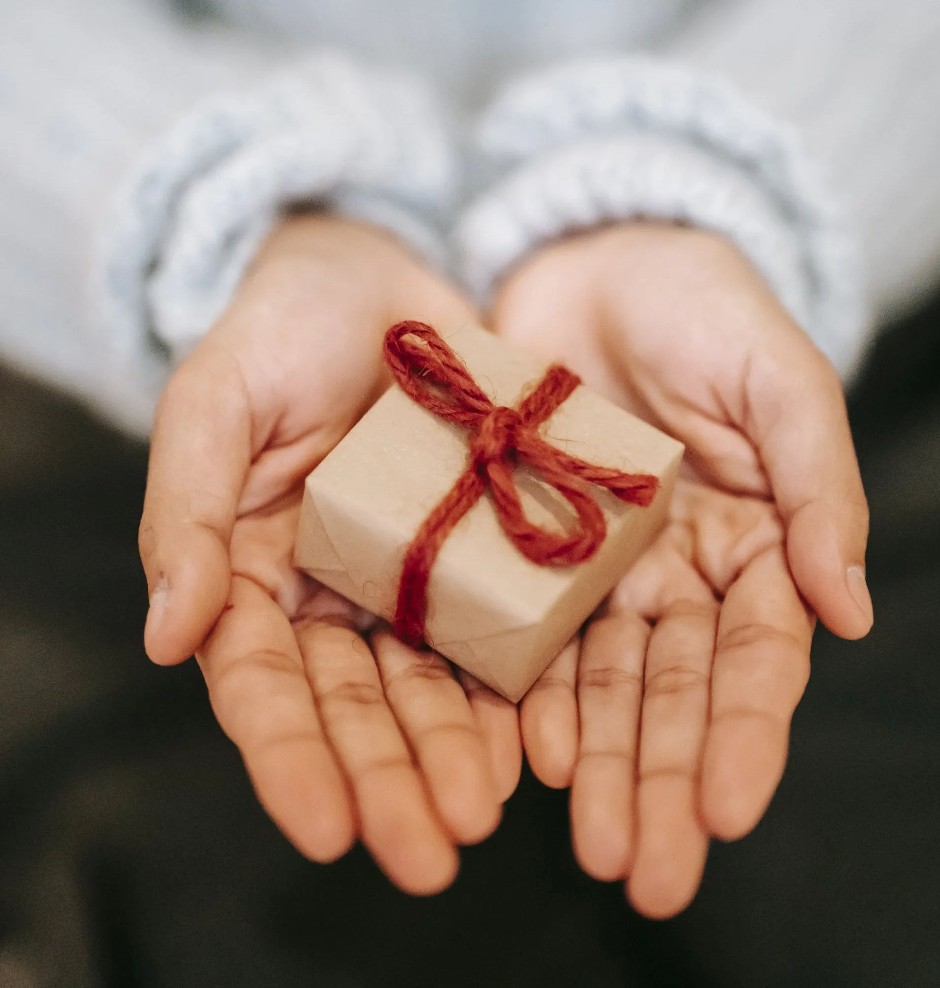 Two hands outstretched holding a small gift wrapped in brown paper and a bow tied with a piece of yarn