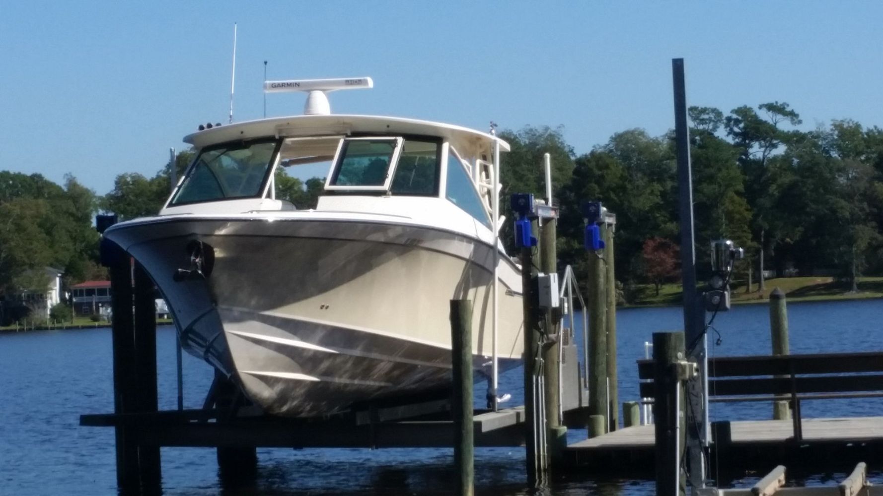 White boat docked at a wooden pier on calm water, with trees and blue sky in the background