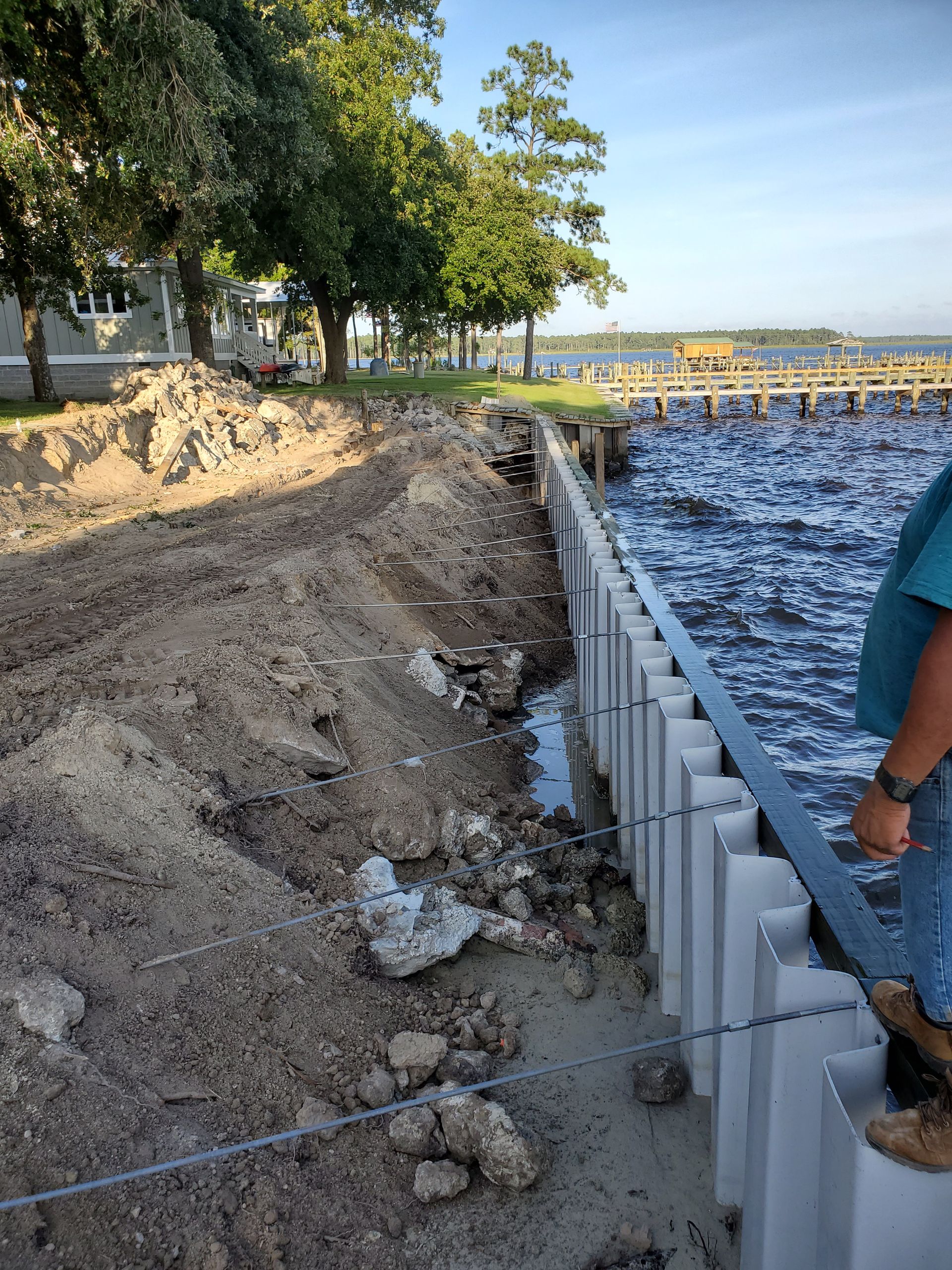 Seawall construction along a sandy shoreline beside trees and calm water, with a person standing at right.