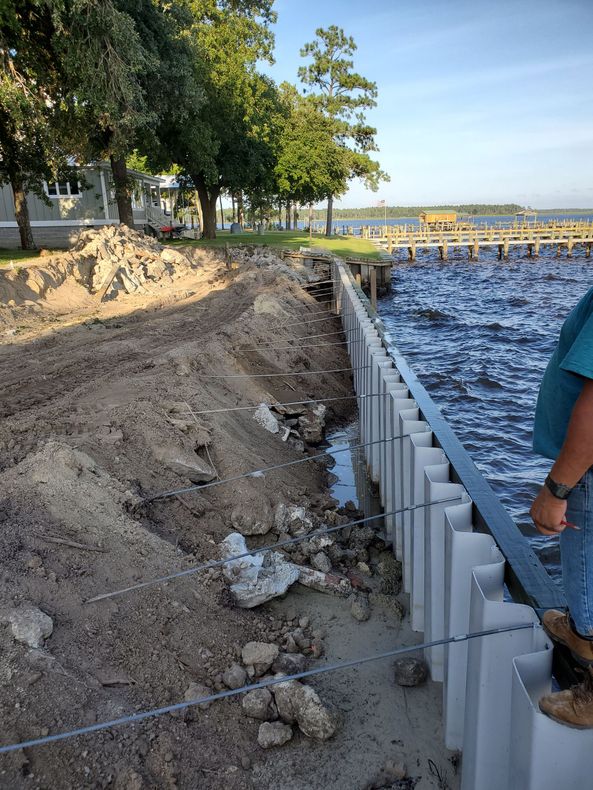 Shoreline repair with stacked white seawall blocks beside water and trees, with a worker at right
