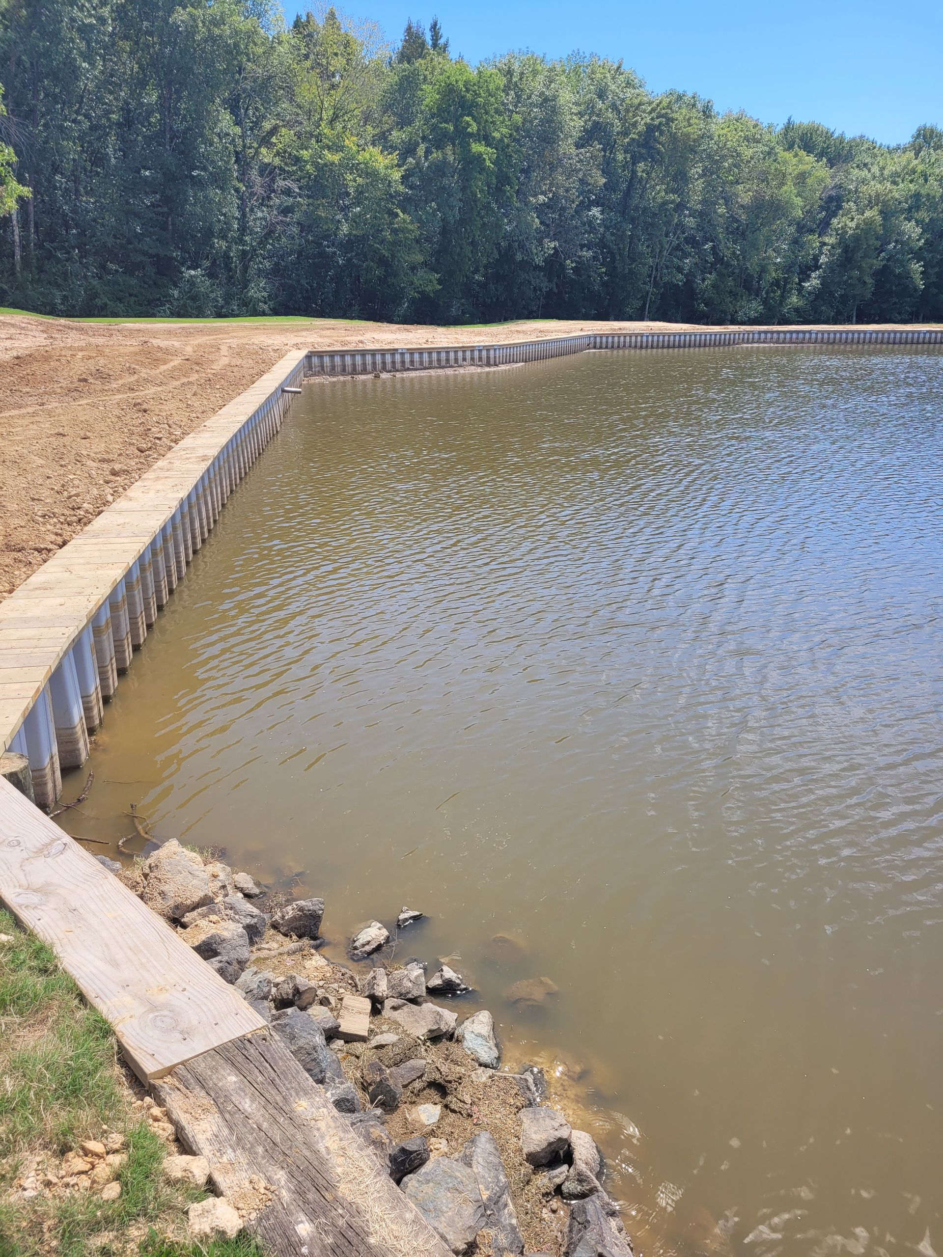 Small concrete-lined pond beside a dirt bank, with muddy water and trees in the background.