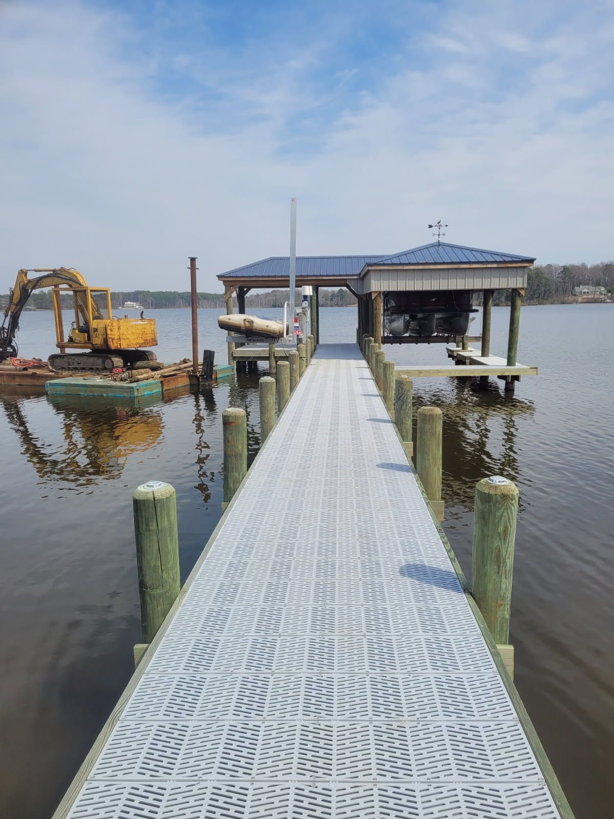 Floating dock leading to a blue-roofed boathouse, with an excavator on the left over calm water.