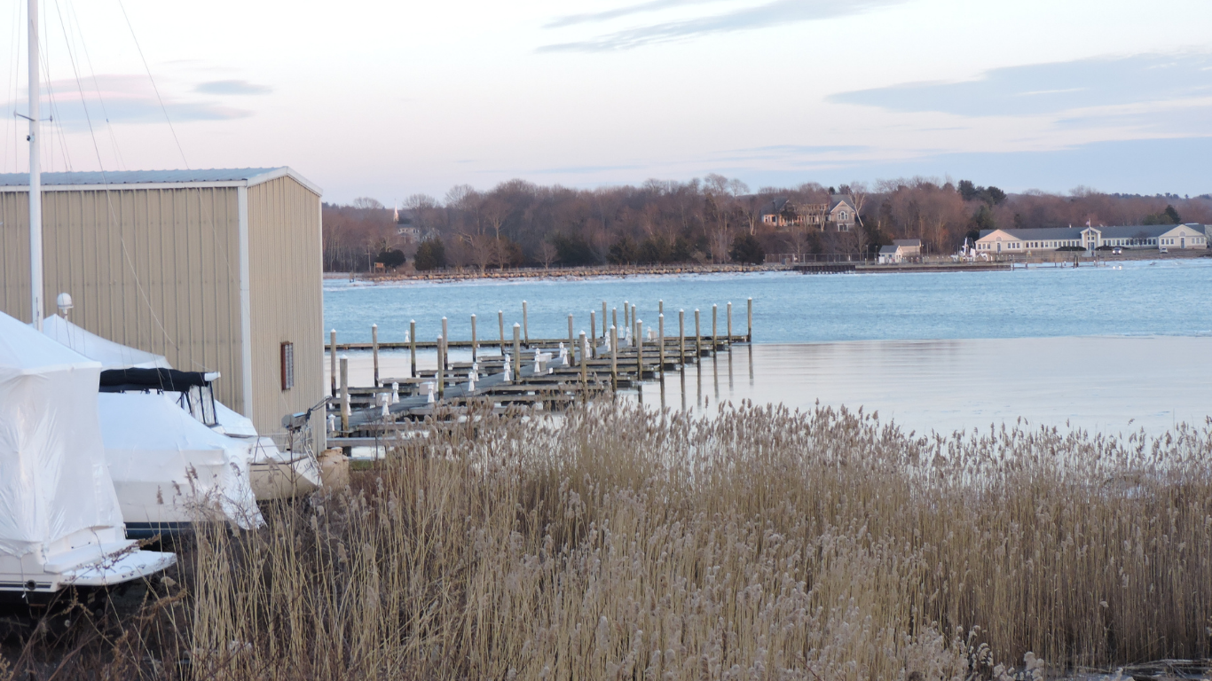 Snowy dock beside a calm lake with reeds and a beige boathouse at left