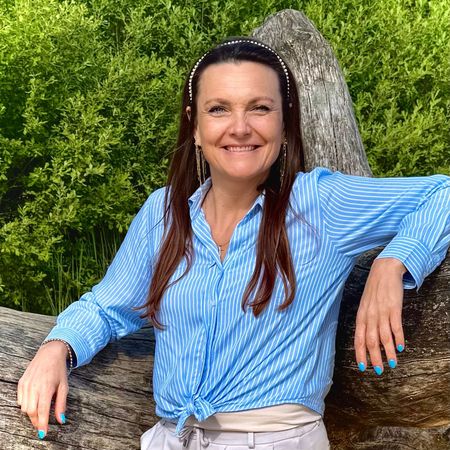 Sabine Een vrouw in een blauw shirt leunt tegen een boomstam.