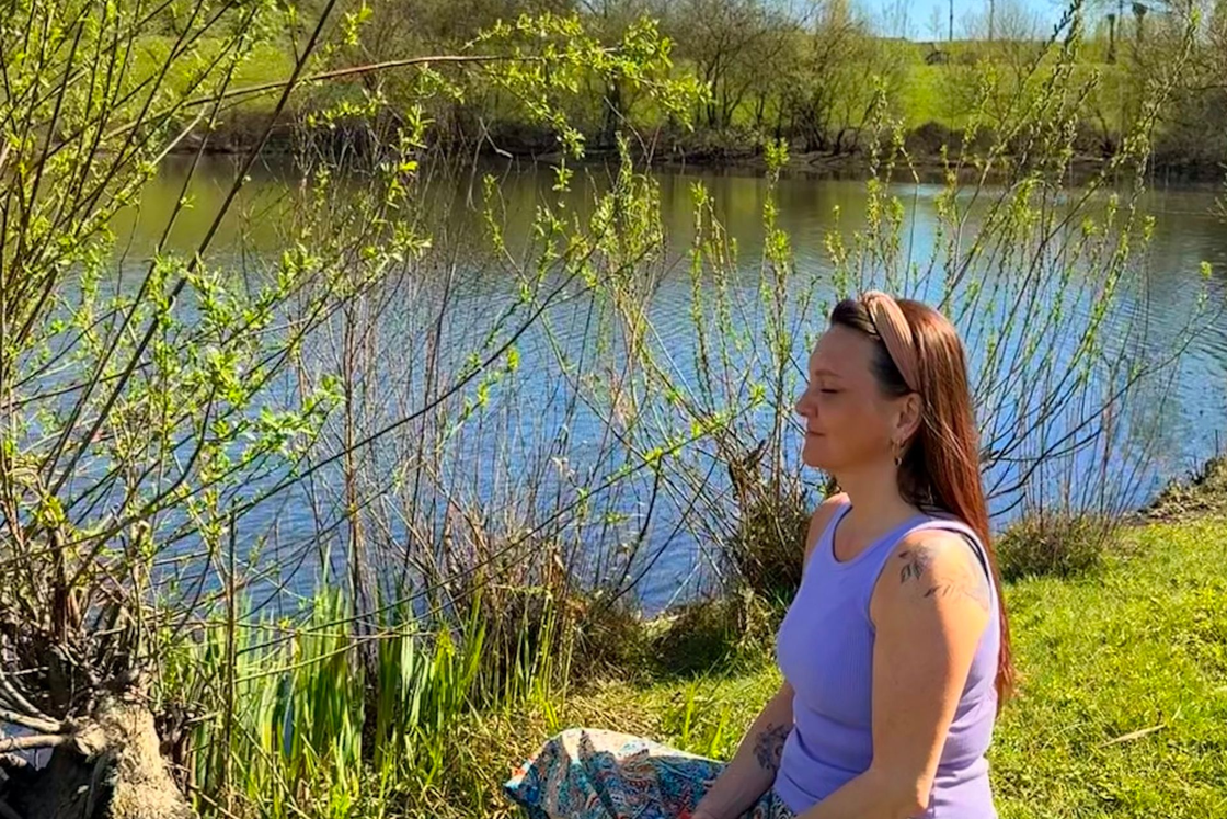 Een persoon zit met gekruiste benen in een meditatieve houding op de grasrijke oever van een vijver onder een helderblauwe hemel.