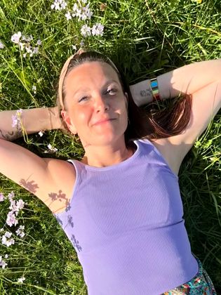 Sabine FHealS in een paarse tanktop ligt in een zonnig, met bloemen gevuld grasveld met de handen achter het hoofd.