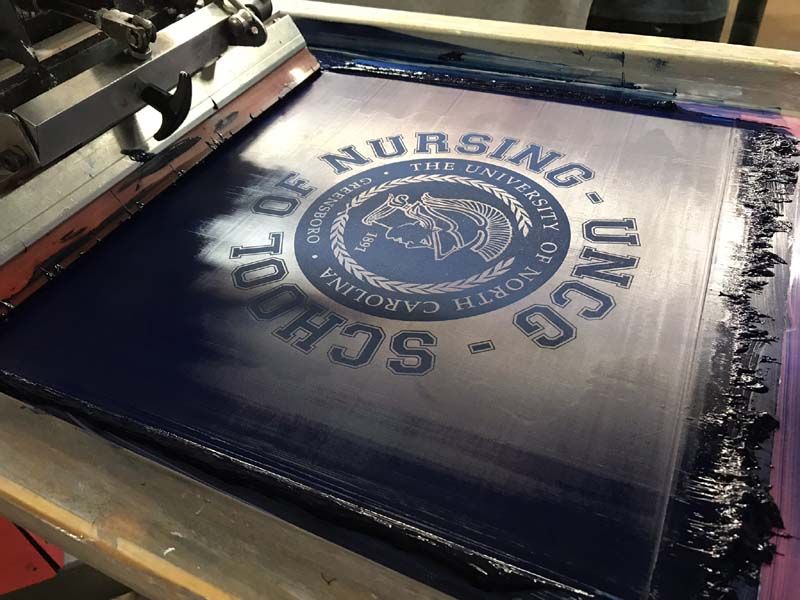 Screen printing a blue UNC Greensboro School of Nursing logo on a navy background.