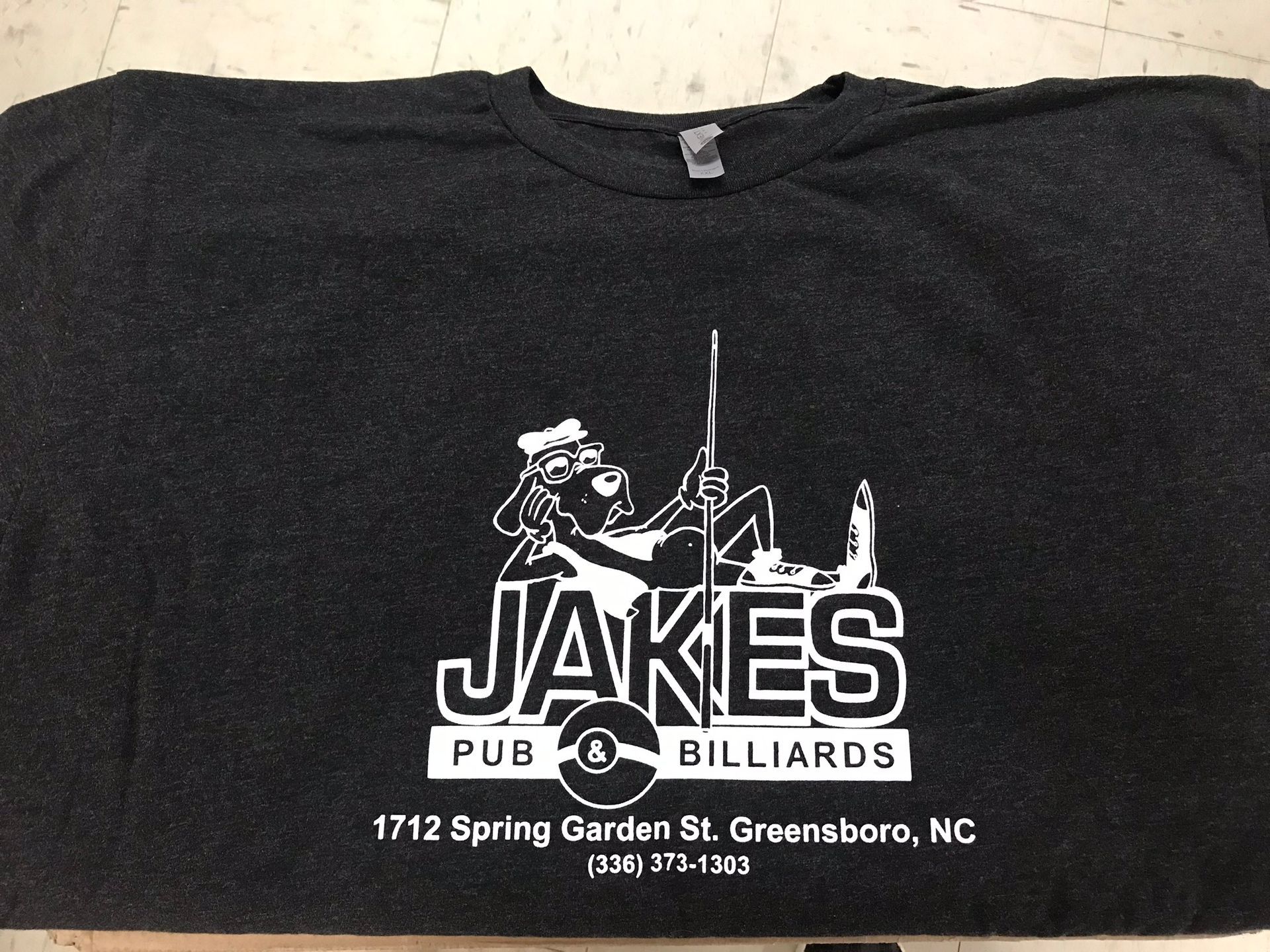 Dark gray t-shirt with a white logo for Jake's Pub & Billiards, Greensboro, NC; a dog-like cartoon character with a pool cue.