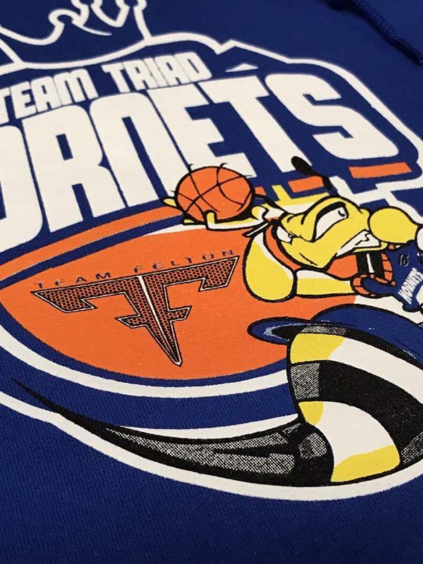 Team Trio Hornets logo on blue fabric. Hornet mascot dribbles basketball, over orange and white stripes.