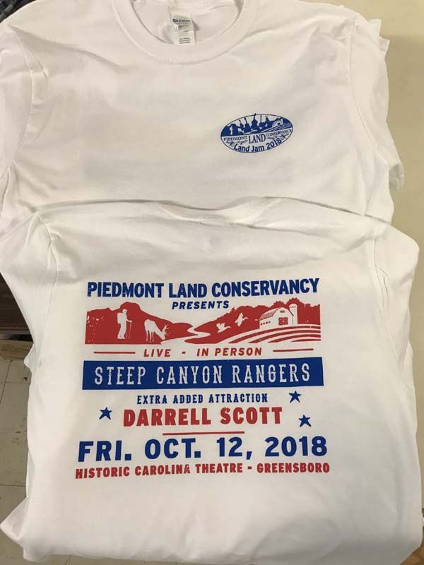 Two white t-shirts with printed logos, one for Piedmont Land Conservancy featuring Steep Canyon Rangers and Darrell Scott.
