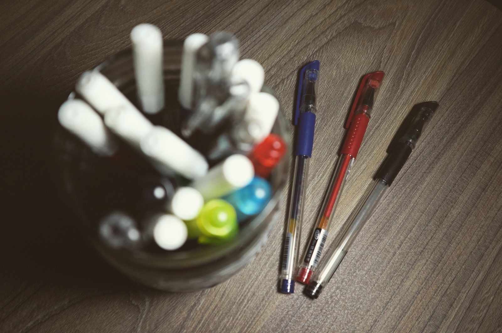 Glass jar filled with pens and three pens in blue, red, and black lying on a wood surface.
