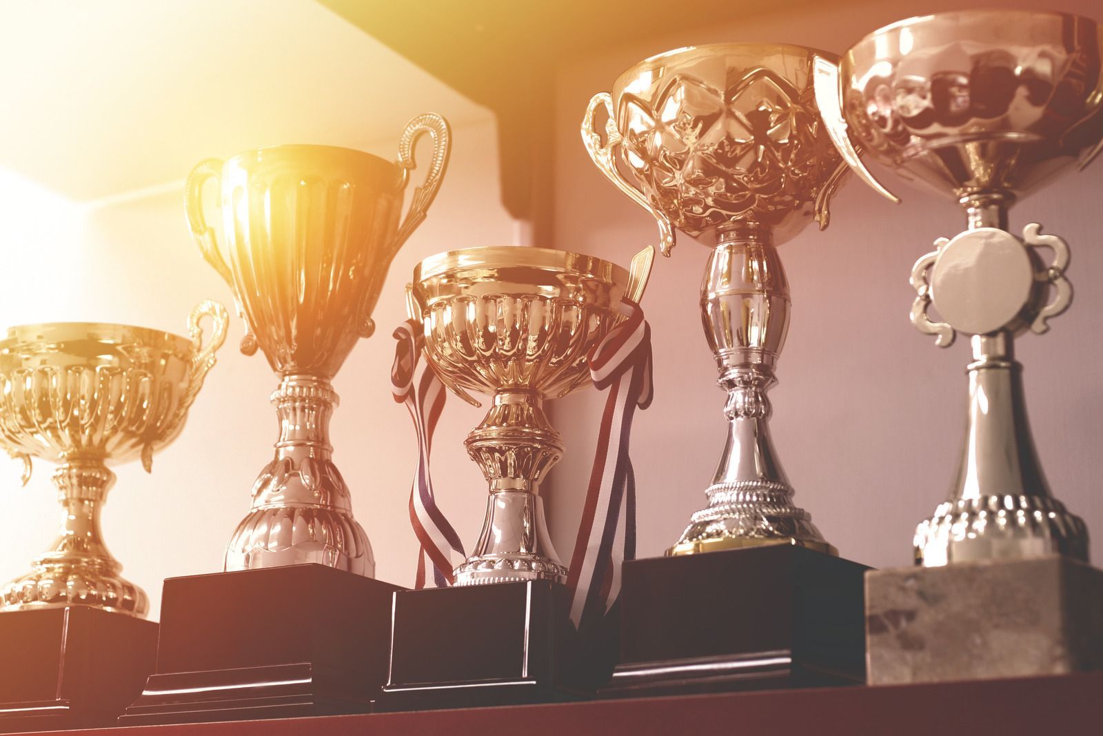 Row of shiny gold trophies on a shelf, bathed in warm light.