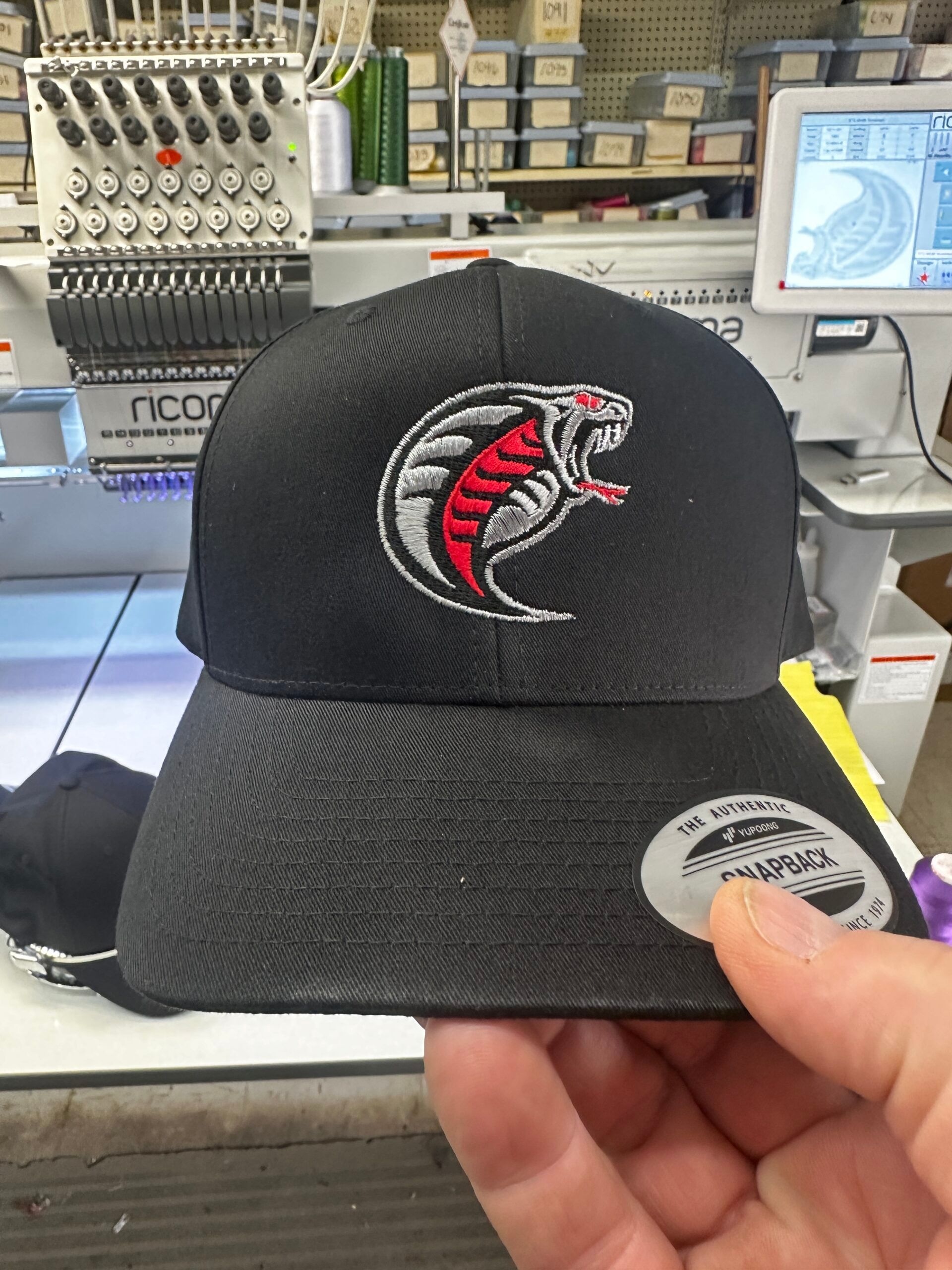 Black baseball cap with cobra logo being held in front of an embroidery machine.