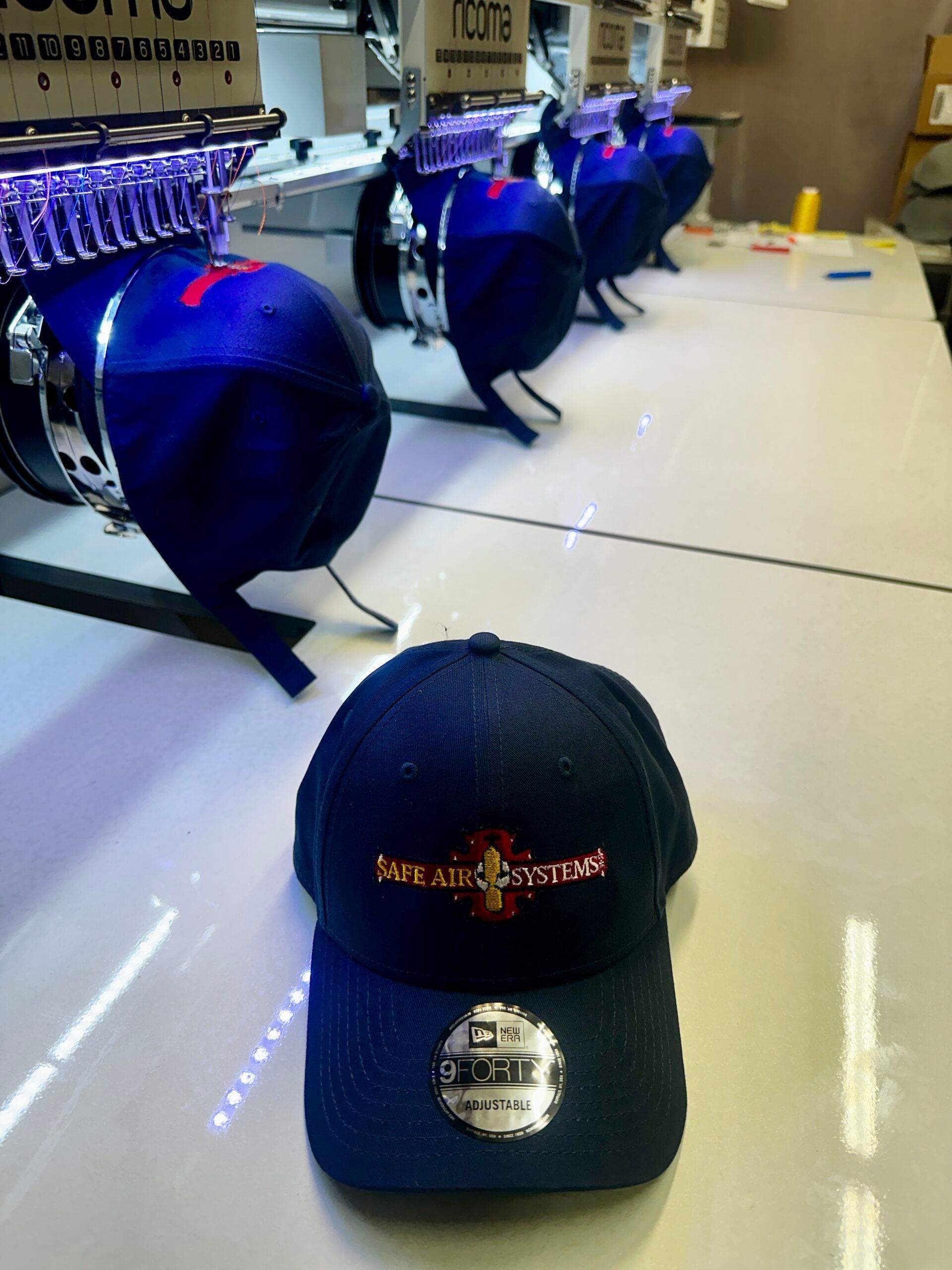 A navy baseball cap with embroidered logo sits in front of embroidery machines.