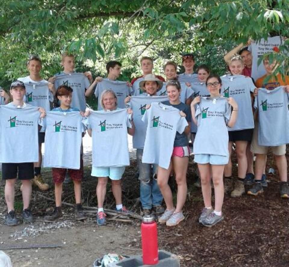 Group holding up matching gray t-shirts, outdoors near foliage. Smiling faces.