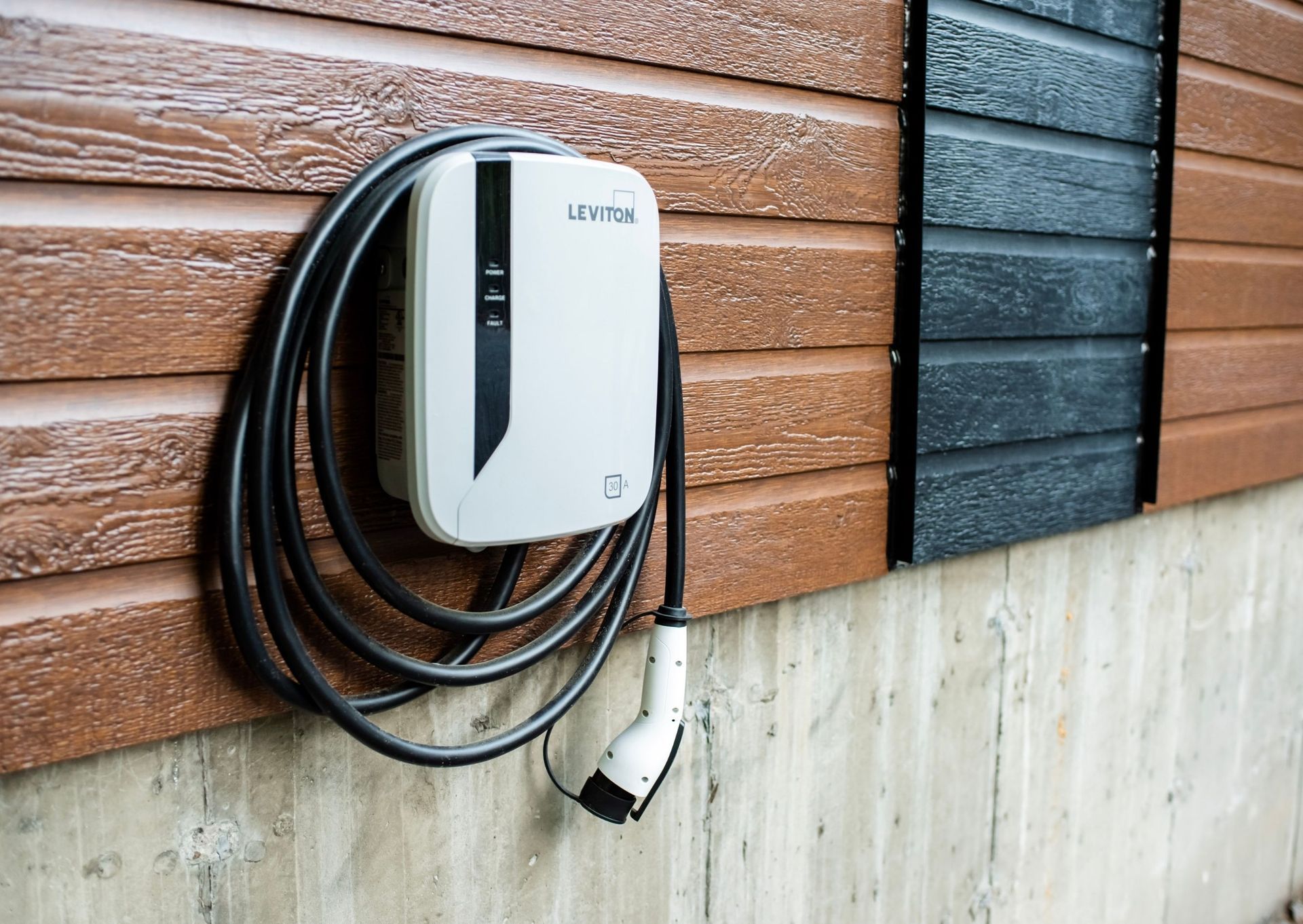An EV charger mounted on a brown and black wood siding wall; the charging cable coiled.