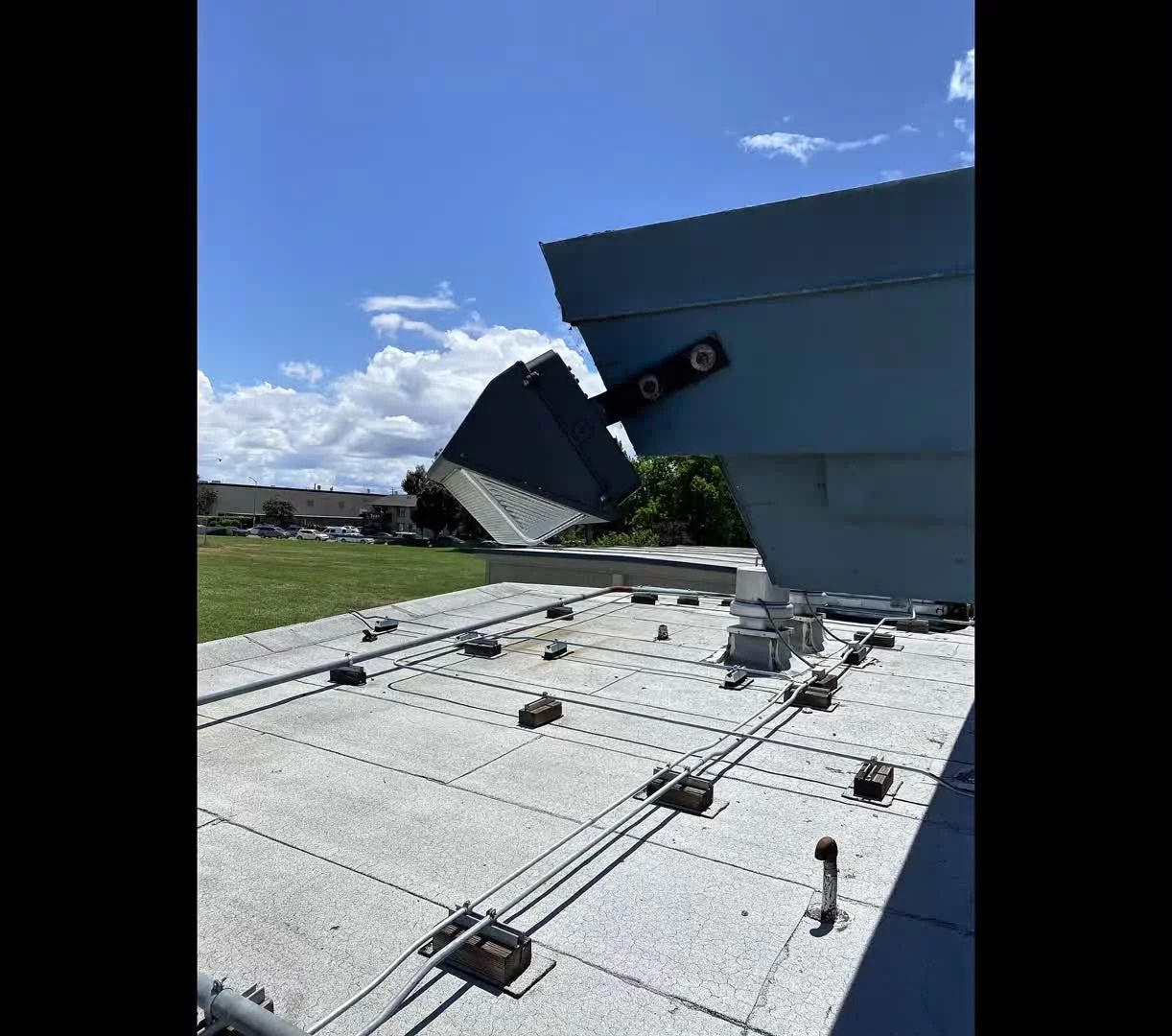 A blue rooftop structure with open metal panels, attached to a flat roof with wiring and fasteners, against a cloudy sky.