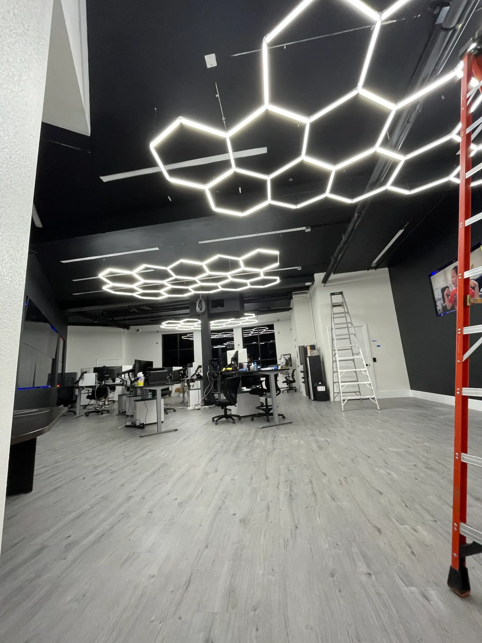 Open office space with black ceiling, hexagonal lights, and workstations; a ladder is in the corner.