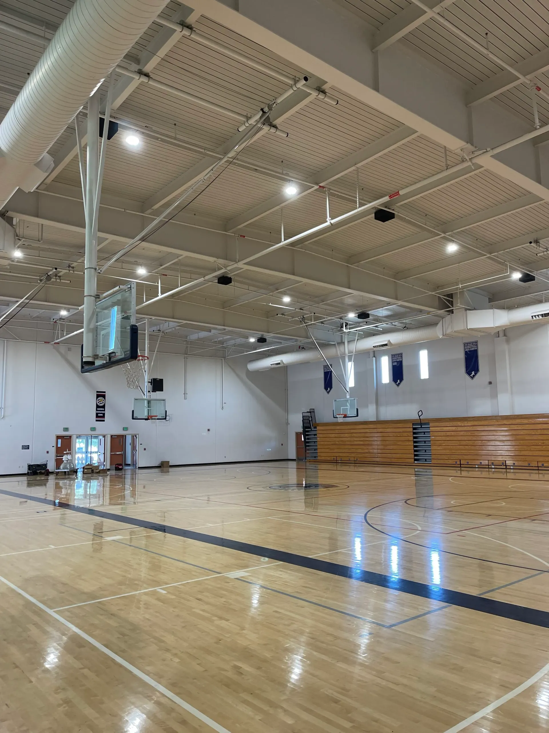 Basketball court interior with hardwood floor, hoops, and retractable bleachers; white ceiling with lights.