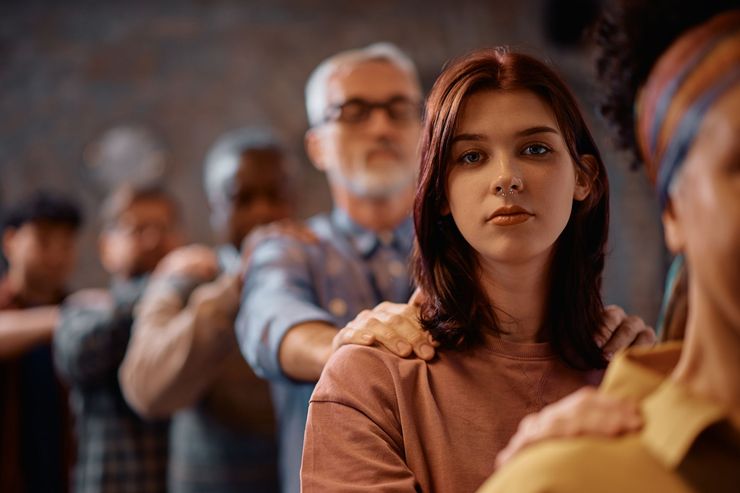 Seated people holding shoulders in a softly lit group setting, with a woman in focus looking forward