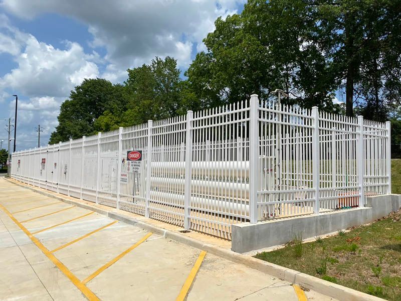 White metal fence around a paved lot under a cloudy sky, with trees in the background.