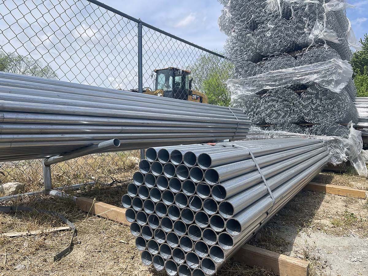 Stack of gray metal pipes beside a chain-link fence, with a forklift in the background.