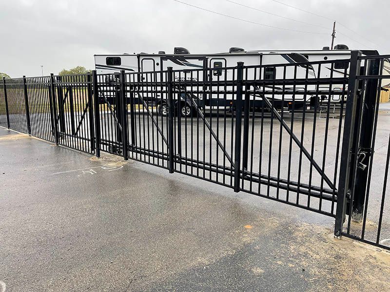 Black metal gate on a wet paved driveway with parked vehicles behind it.