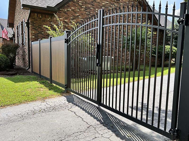 Black metal driveway gate beside a brick house and fence on a sunny residential street
